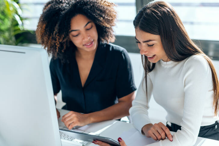 Two ladies sat at a desk in front of a computer looking at an iPad